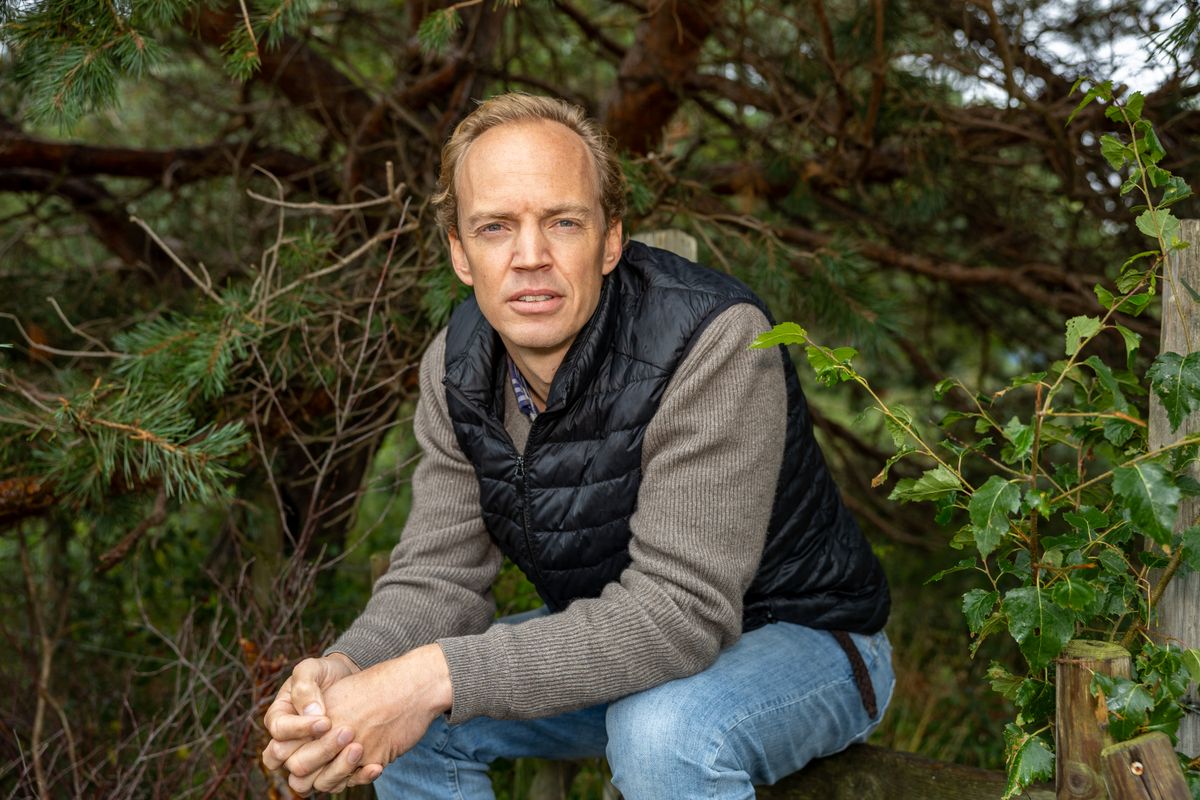 Man i svart väst och grå tröja sitter utomhus framför täta gröna trädgrenar