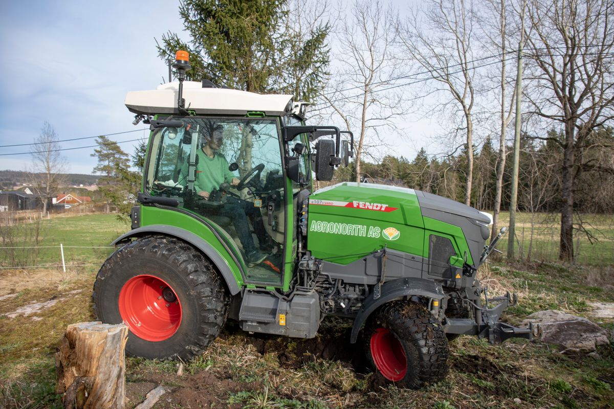 Grön traktor med förare kör på ojämn mark i lantlig miljö nära skog och åker.