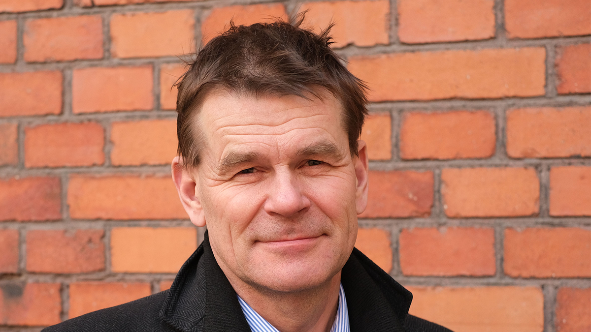Leende medelålders man i mörk kappa framför en tegelvägg