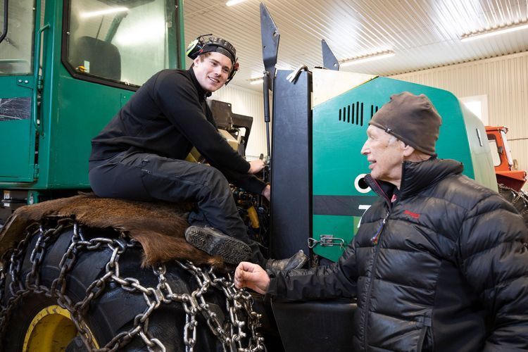 Ung man på skogsmaskin med kedjor på däcken pratar med äldre man i verkstad